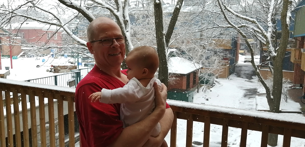 Photo of Papa Zesser holding Asta in his arms, while standing on his back porch as late-March snow falls from the sky.
