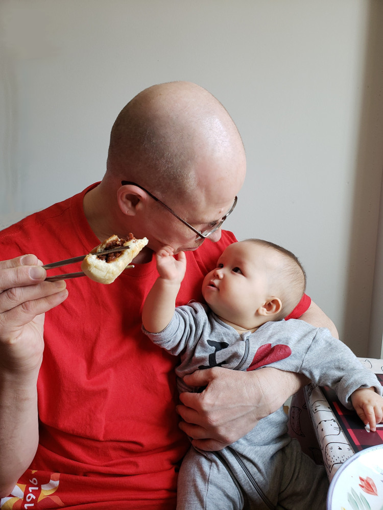 Photo of Papa Zesser holding a partially-eaten bar-b-q pork bun in chopsticks, while holding Baobao in his left arm as she reaches for his chin.