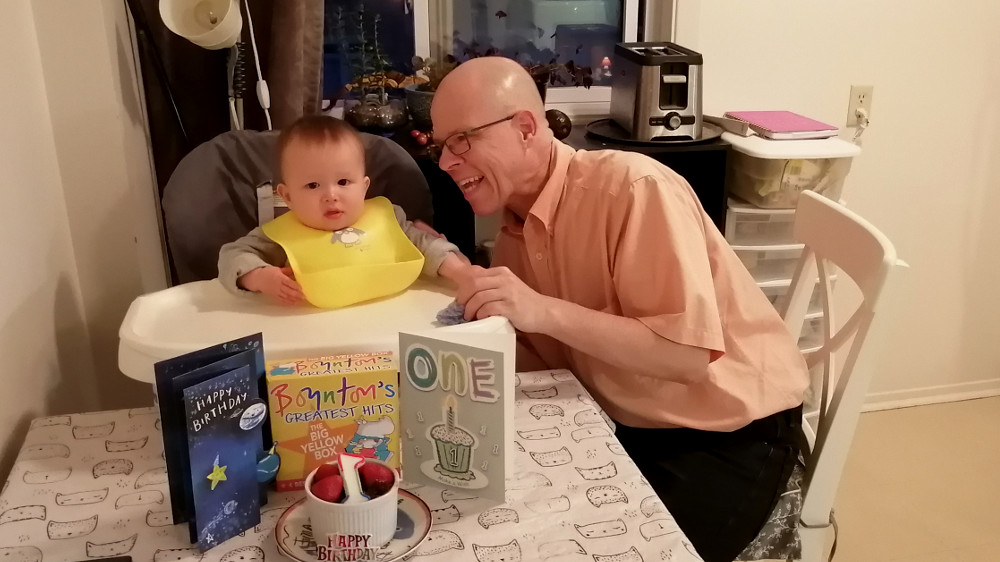 Photo of Papa Zesser with Baby Baobao at table with cards and cake for first birthday, August 19, 2020.