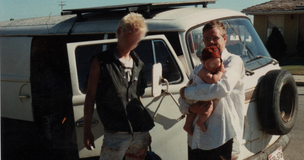 Photo of a very young Papa Zesser holding a friend's baby in his arms while posing, with another friend, in front of his 1971 Volkswagen microbus.