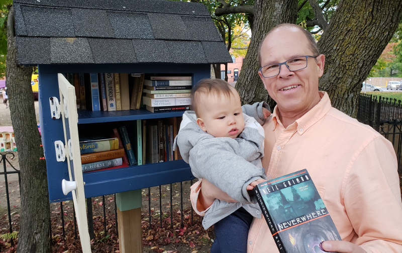 Photo of Papa Zesser holding baby Baobao in front of local little library, about to return Neil Gaiman's not-so-good book, Neverwhere.