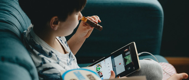 Detail of a photo of a child seated on a chair or couch, holding a tablet in one hand and a half-eaten chocolate bar in the other. Image via Unsplash.com by @anniespratt.